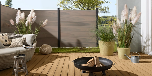 Runde Sonnenliege auf heller WPC-Terrasse mit Ziergräsern und stilvoller Feuerstelle. Im Hintergrund WPC-Zaun