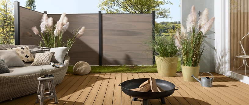 Runde Sonnenliege auf heller WPC-Terrasse mit Ziergräsern und stilvoller Feuerstelle. Im Hintergrund WPC-Zaun