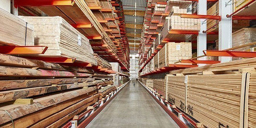 Großes Holzlager mit gestapeltem Schnittholz, Kanthölzern und Brettern in hohen Regalen mit roten Trägern.